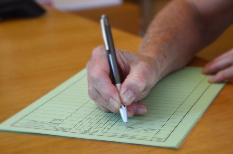 Eine ältere Person schreibt mit einem Stift auf einem Holztisch auf ein Formular. Das Formular hat mehrere Zeilen.