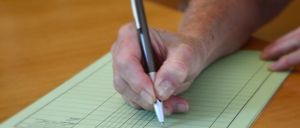 Eine ältere Person schreibt mit einem Stift auf einem Holztisch auf ein Formular. Das Formular hat mehrere Zeilen.