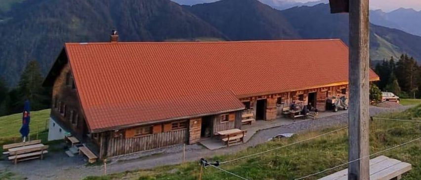 Ein Holzkreuz steht neben einem rotgedeckten Chalet vor einer Bergkulisse. Das Chalet hat Bänke und einen geschlossenen Schirm in der Nähe. Der Himmel ist klar mit einigen Wolken.