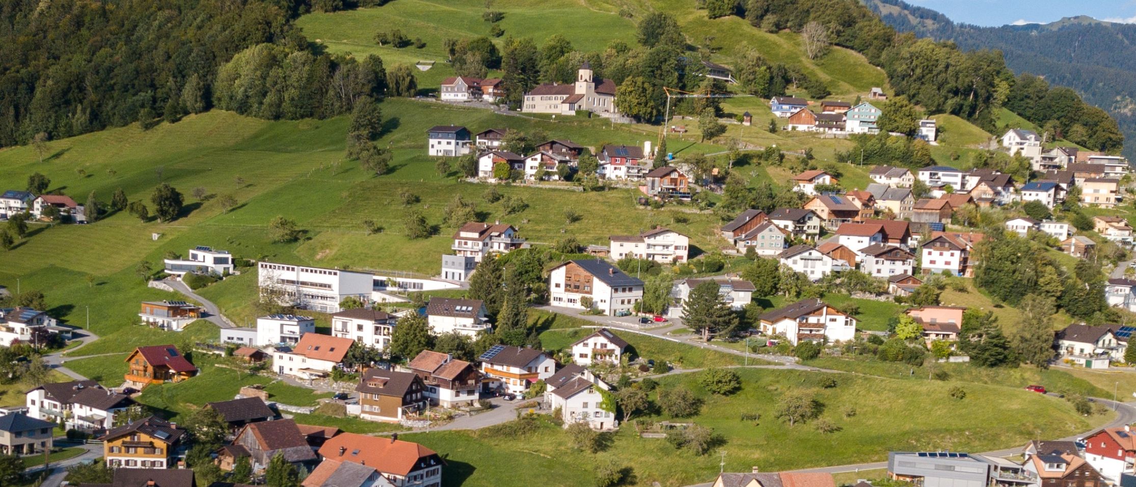 Luftaufnahme eines Dorfes am Hang mit grünen Wiesen und Bäumen, Häusern in verschiedenen Farben und Größen sowie einer kurvenreichen Straße.