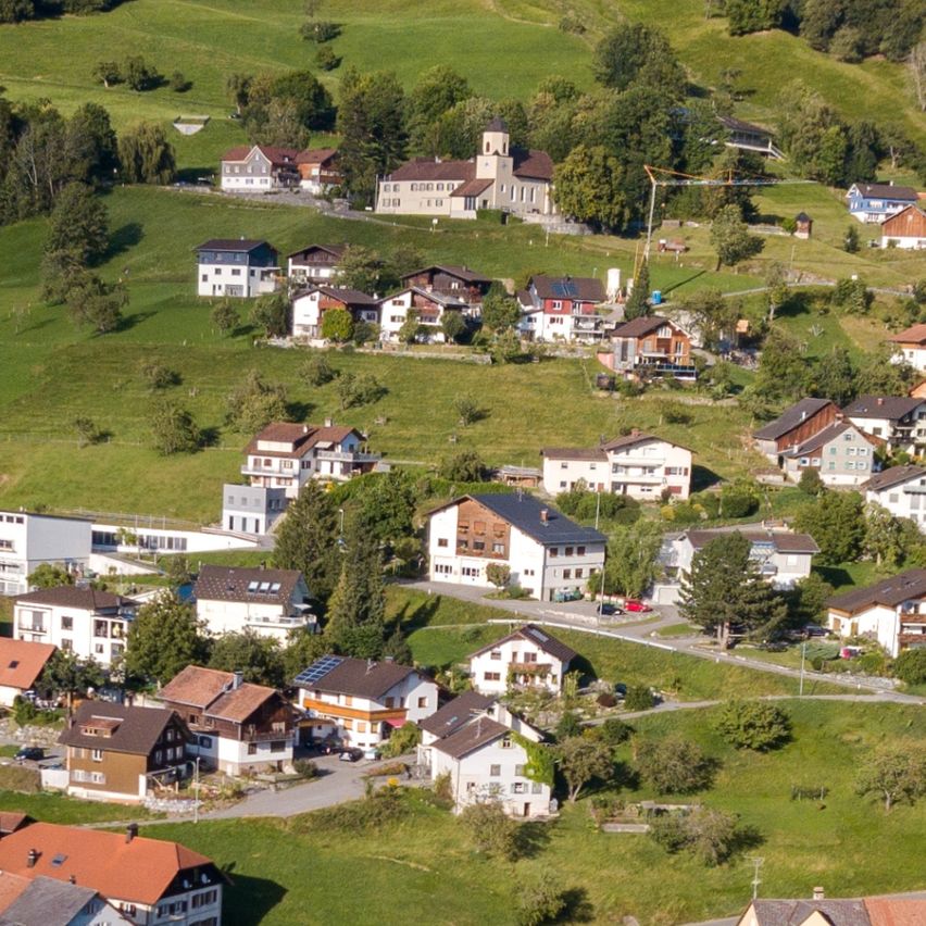 Luftaufnahme eines Dorfes am Hang mit grünen Wiesen und Bäumen, Häusern in verschiedenen Farben und Größen sowie einer kurvenreichen Straße.