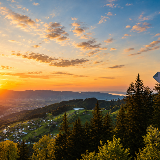 Ein Panoramablick auf einen Sonnenuntergang über einem Tal mit Bergen in der Ferne, einem Dorf unten und einem klaren blauen Himmel mit verstreuten Wolken.