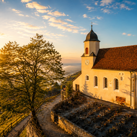 Eine kleine Kirche mit einem Turm und einer Uhr befindet sich auf einem Hügel, umgeben von einem Friedhof und einem großen Baum. Der Himmel ist blau mit Wolken.