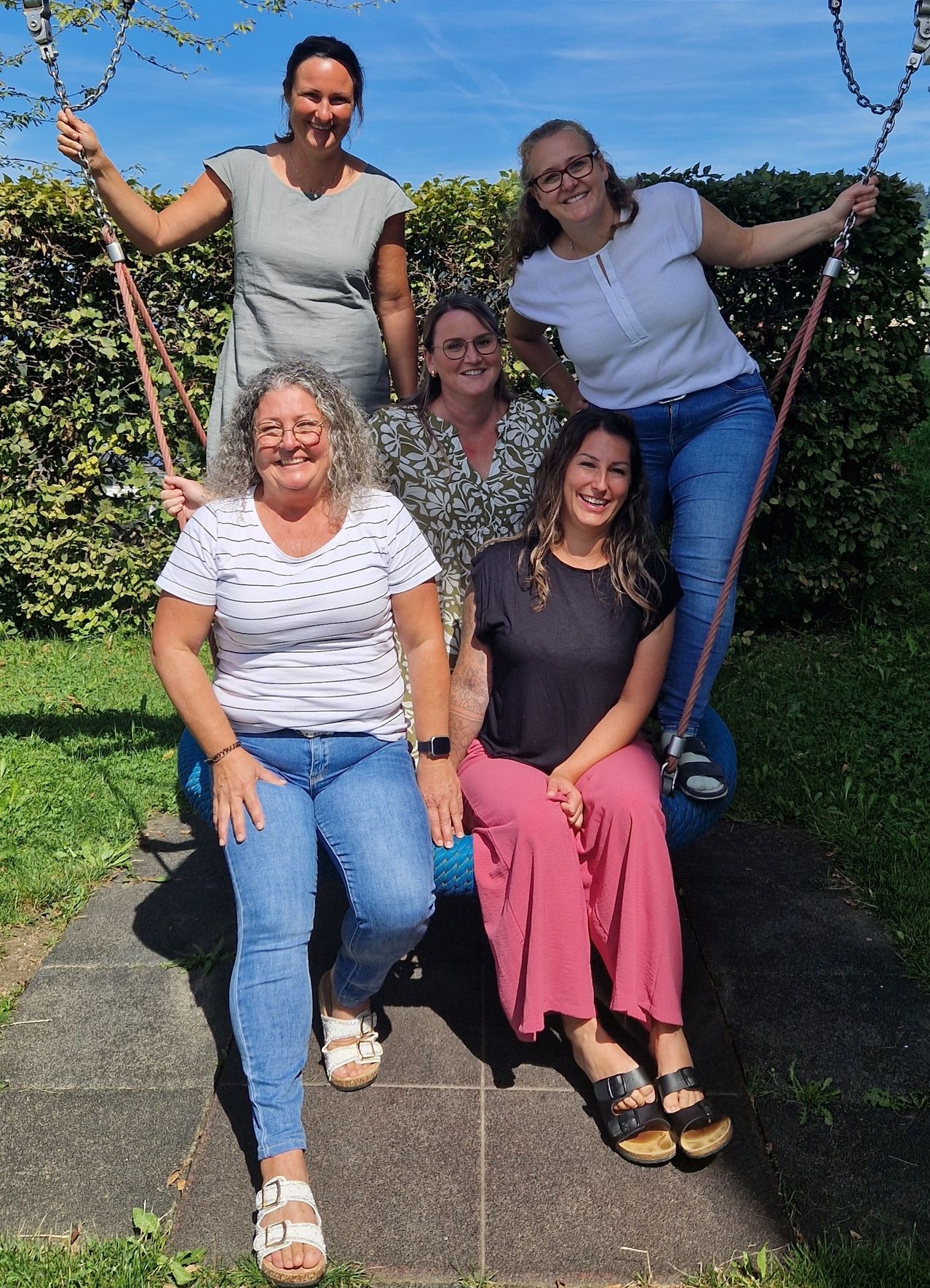 Fünf Frauen, lächelnd, posieren zusammen auf einer Schaukel und einem blauen Fitnessball in einem Garten mit grünen Büschen im Hintergrund.