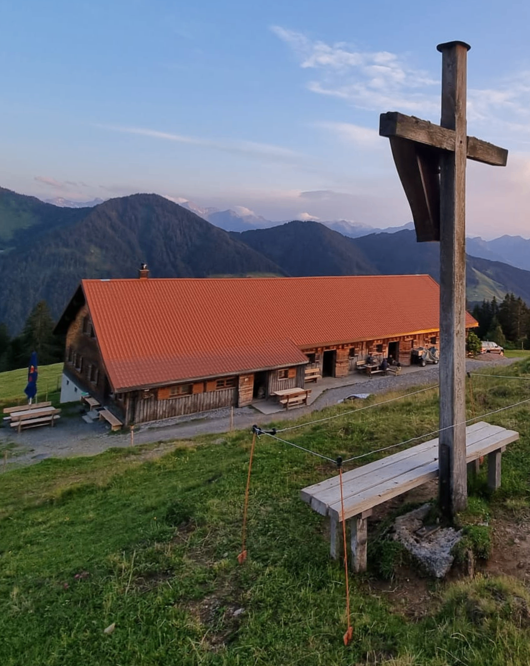 Ein rustikales Holzhaus mit rotem Dach steht auf einem Grashügel mit einem Holzkreuz und einer Bank im Vordergrund, umgeben von Bergen und einem klaren Himmel.