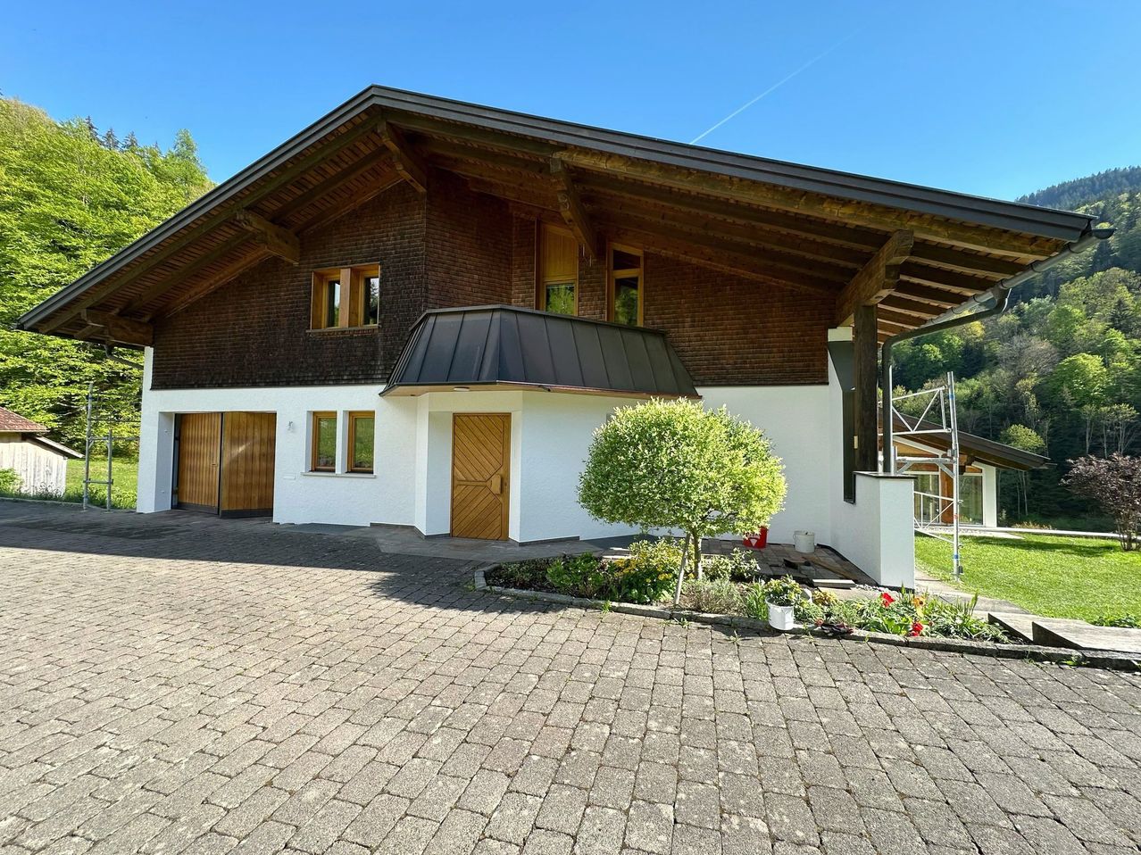 Ein modernes Chalet mit brauner Holzfassade und weißen Wänden, in einer malerischen Gegend mit grünen Bäumen und einem klaren blauen Himmel.