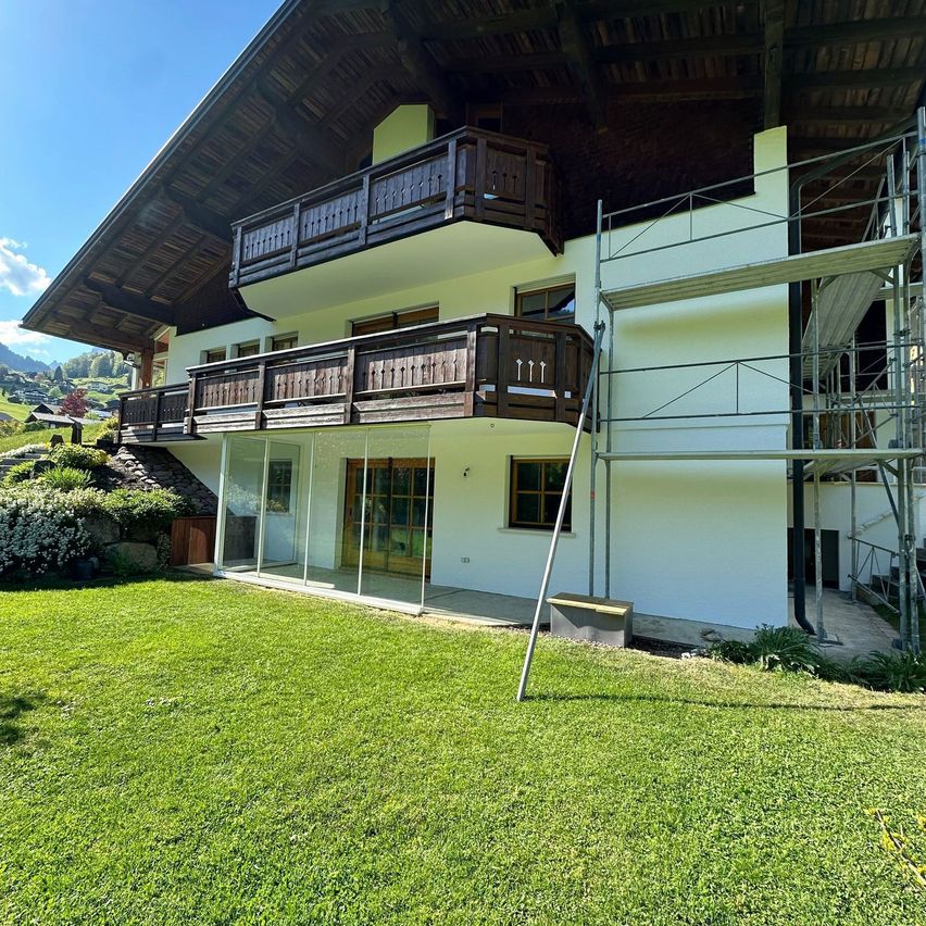 Ein Haus mit Balkon und Garten wird gebaut. Das Haus hat eine Glastür und Glasfenster. Das Haus hat ein schräges Dach und einen Schornstein.