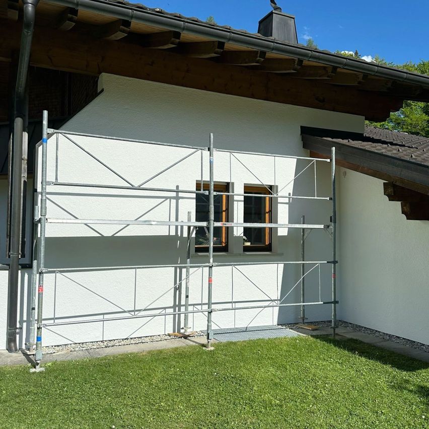 Ein Gerüst ist vor einem weißen Haus mit einem braunen Dach aufgestellt. Das Haus hat zwei Fenster und einen Schornstein.