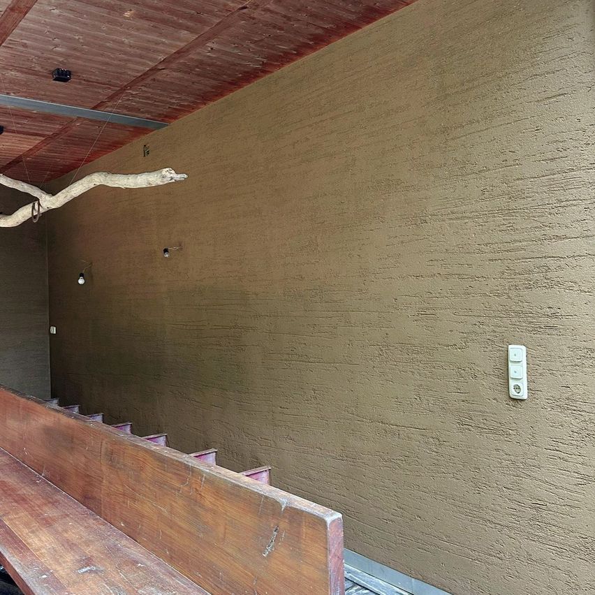 Ein Innenraum mit einer strukturierten Wand und einer Holzbank, ein Ast hängt von der Decke mit kleinen Lichtern. Es gibt einen weißen Schalter an der Wand.