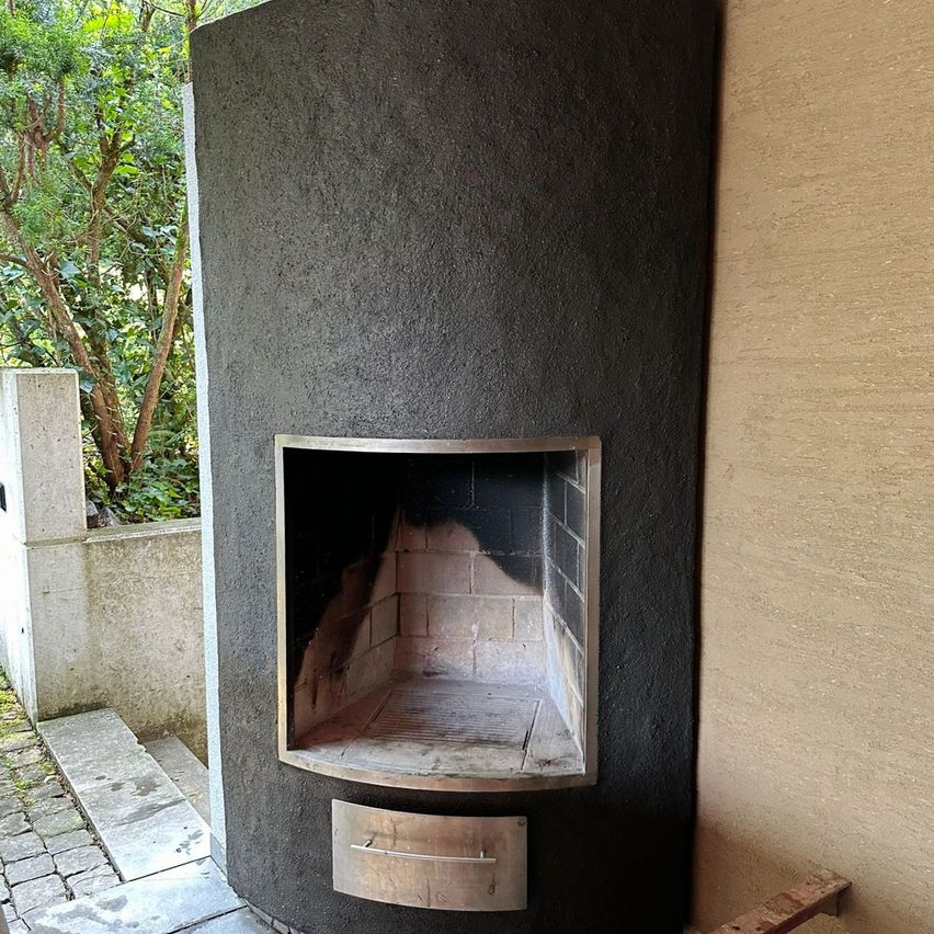 Ein Außenkamin ist in eine Betonwand integriert. Der Kamin hat eine rechteckige Öffnung mit Metallrahmen und Ziegelinnenseite. Ein Holzbrett befindet sich in der Nähe.