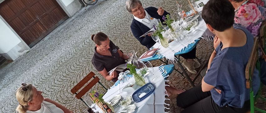 Eine Gruppe von Menschen sitzt um Tische auf einer Kopfsteinpflasterstraße, möglicherweise beim Essen. Flaschen, Gläser und Pflanzen stehen auf den Tischen. Hinter der Gruppe sind Fahrräder geparkt.