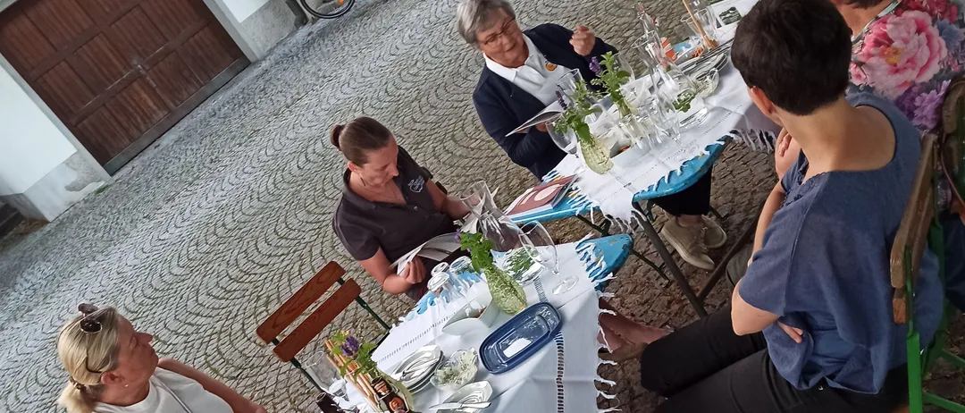 Eine Gruppe von Menschen sitzt um Tische auf einer Kopfsteinpflasterstraße, möglicherweise beim Essen. Flaschen, Gläser und Pflanzen stehen auf den Tischen. Hinter der Gruppe sind Fahrräder geparkt.