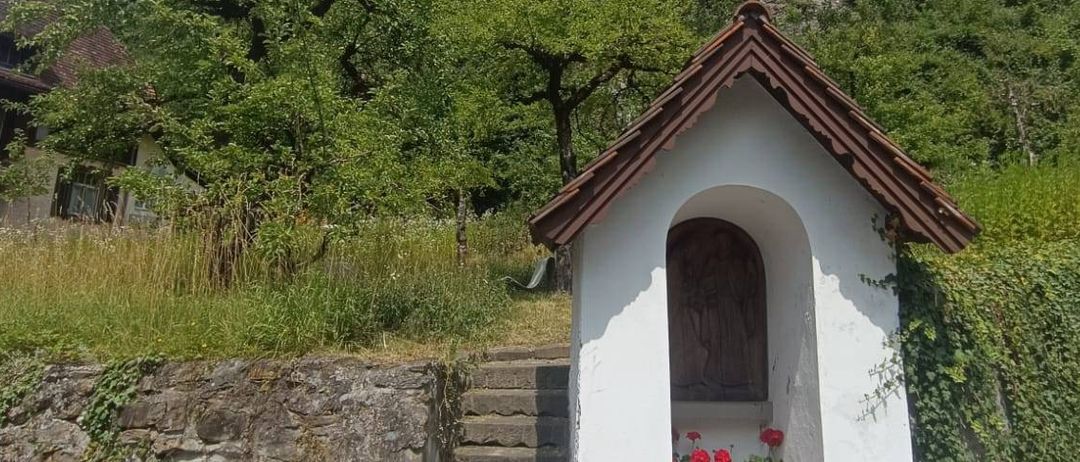 Eine kleine weiße Kapelle mit einem braunen Dach steht an der Straßenseite, mit Stufen, die zu ihr hinaufführen. Sie hat eine Holztür und Blumen davor. Dahinter steht ein Turm auf einem Felsen mit einer Uhr darauf.