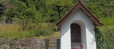 Eine kleine weiße Kapelle mit einem braunen Dach steht an der Straßenseite, mit Stufen, die zu ihr hinaufführen. Sie hat eine Holztür und Blumen davor. Dahinter steht ein Turm auf einem Felsen mit einer Uhr darauf.