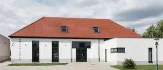 Ein modernes weißes Gebäude mit rotem Dach, mehreren Fenstern und einer Einfahrt davor. Es hat eine Straßenlampe und etwas Grün.