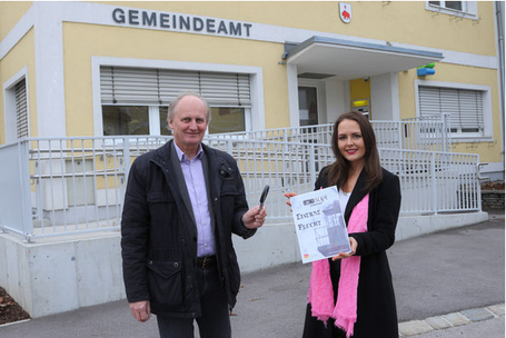 Ein Mann und eine Frau stehen vor einem Gebäude mit der Aufschrift 'Gemeindeamt'. Der Mann hält einen Stift und Papier, während die Frau ein weißes Papier mit schwarzem Umriss hält.