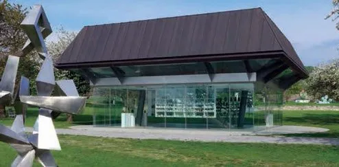 Ein modernes Gebäude mit einem braunen Dach und Glaswänden befindet sich in einem Park mit einem Rasenfeld. Es verfügt über eine große abstrakte Metallskulptur vor der Fassade.