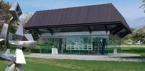 Ein modernes Gebäude mit einem braunen Dach und Glaswänden befindet sich in einem Park mit einem Rasenfeld. Es verfügt über eine große abstrakte Metallskulptur vor der Fassade.