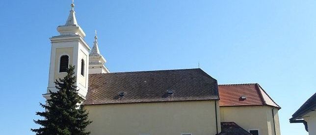 Eine große weiße Kirche mit einem braunen Dach und zwei weißen Türmen steht im Vordergrund. Ein hoher Kiefernbaum steht davor, mit einer weißen Einzäunung und verschiedenen Pflanzen rund um das Gebäude.