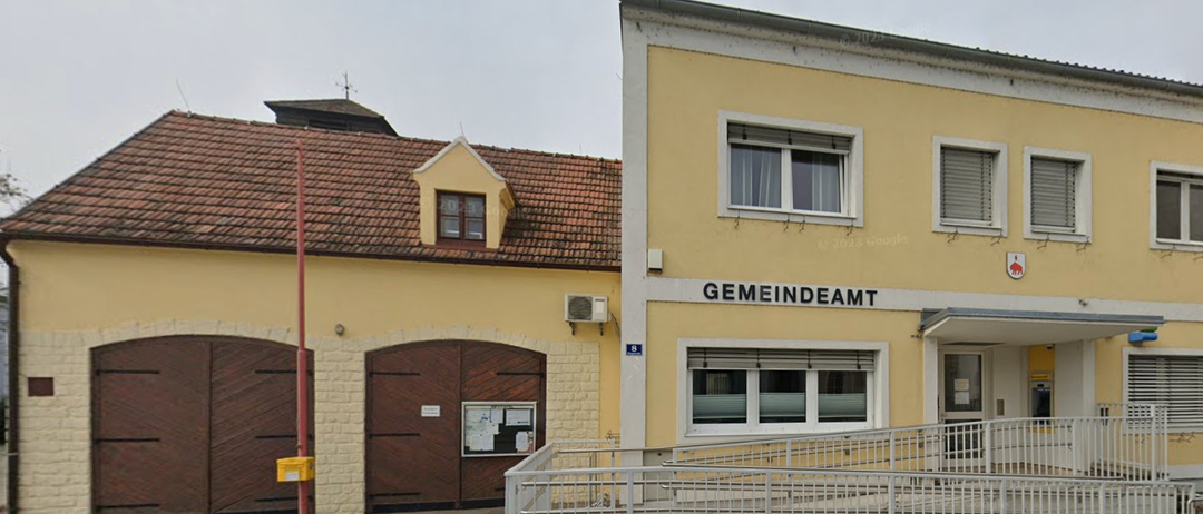 Ein gelbes Gebäude mit braunem Dach, Fenstern und Türen. Das Gebäude trägt die Aufschrift 'GEMEINDEAM'. Eine Klimaanlage ist an der Wand angebracht. Rechts befindet sich eine Treppe.
