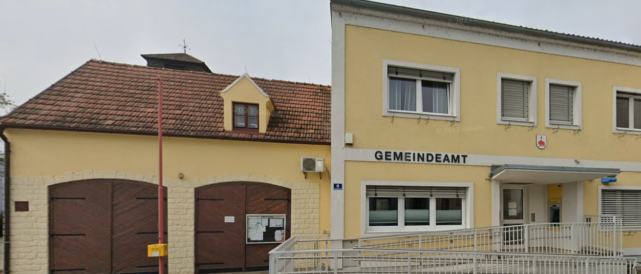 Ein gelbes Gebäude mit braunem Dach, Fenstern und Türen. Das Gebäude trägt die Aufschrift 'GEMEINDEAM'. Eine Klimaanlage ist an der Wand angebracht. Rechts befindet sich eine Treppe.