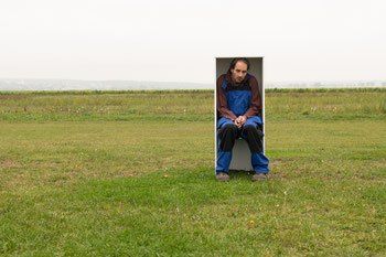 Ein Mann in einem blauen Overall sitzt in einem weißen Kasten in einem großen grünen Feld unter einem bewölkten Himmel.