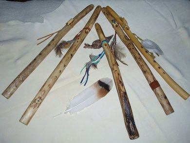 Sechs hölzerne Flöten, kunstvoll geschnitzt und mit Federn verziert, sind auf einem weißen Tuch ausgestellt. Jede Flöte zeigt einzigartige Designs und Federn, was auf kulturelle oder zeremonielle Bedeutung hindeutet.
