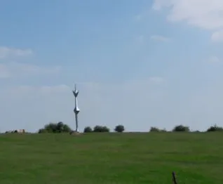 Eine hohe Metallstruktur steht in der Mitte eines weitläufigen, grünen Feldes unter einem teilweise bewölkten Himmel.