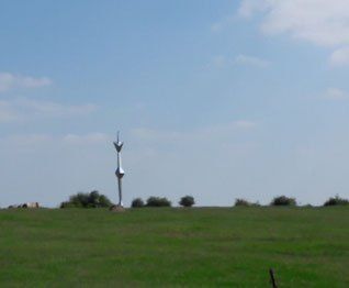 Eine hohe Metallstruktur steht in der Mitte eines weitläufigen, grünen Feldes unter einem teilweise bewölkten Himmel.