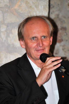 Ein Mann in einem schwarzen Anzug, der ein Mikrofon hält und bei einer Veranstaltung spricht. Er steht vor einer Steinmauer.