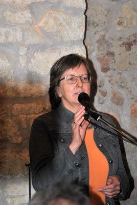 Eine Frau mit Brille und einem orangefarbenen Oberteil spricht in ein Mikrofon und steht vor einer Steinmauer.