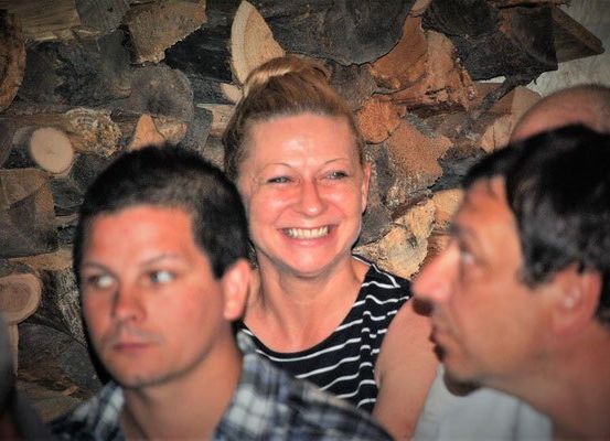Eine Frau mit lächelndem Gesicht steht zwischen zwei Männern. Sie hat eine Duttfrisur. Sie sind wahrscheinlich in einem Raum mit einer holzvertäfelten Wand.