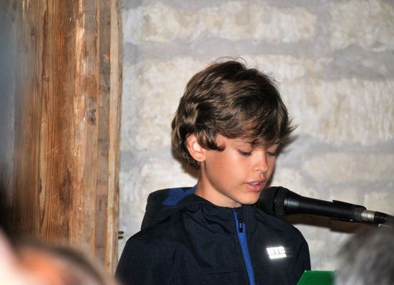 Ein junger Junge in einem schwarzen Mantel mit blauem Reißverschluss spricht in ein Mikrofon, mit einer Ziegelwand und einem Holzpfosten hinter ihm.