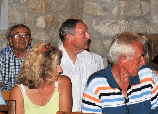 Eine Gruppe älterer Erwachsener sitzt in einem Raum, möglicherweise bei einer Besprechung. Der Mann im weißen Hemd schaut etwas an. Die Frau trägt eine Sonnenbrille auf dem Kopf.