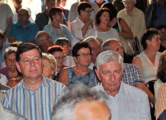 Eine Menschenmenge sitzt und steht in einem großen Saal, möglicherweise bei einer Veranstaltung. Einige tragen Brillen, und eine Frau hat die Hand im Gesicht.