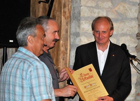 Drei Männer stehen neben einem Podium, einer hält ein gelbes Zertifikat, der andere lächelt. Sie stehen vor einer Ziegelsteinwand und einem Mikrofon.