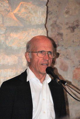 Ein älterer Mann mit Brille und einem schwarzen Blazer spricht in ein Mikrofon bei einer Veranstaltung.