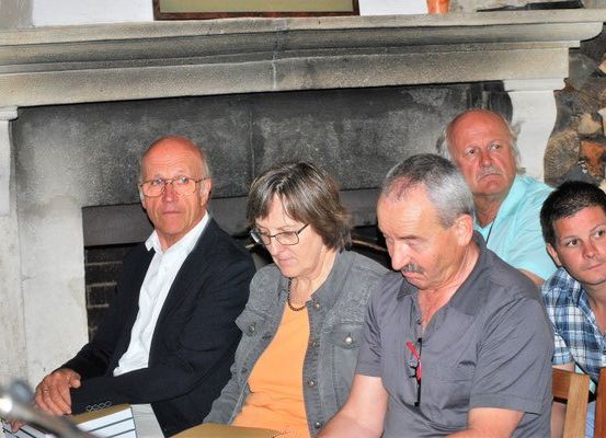 Fünf Erwachsene sitzen in einem Raum, drei Männer und zwei Frauen, wobei eine Bücher hält. Dahinter befindet sich ein Kamin mit einer Steinmauer und einem Bilderrahmen darüber.