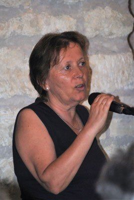Eine Frau mit kurzen Haaren spricht in ein Mikrofon. Sie trägt ein schwarzes ärmelloses Oberteil und eine Halskette. Hinter ihr befindet sich eine Steinmauer.