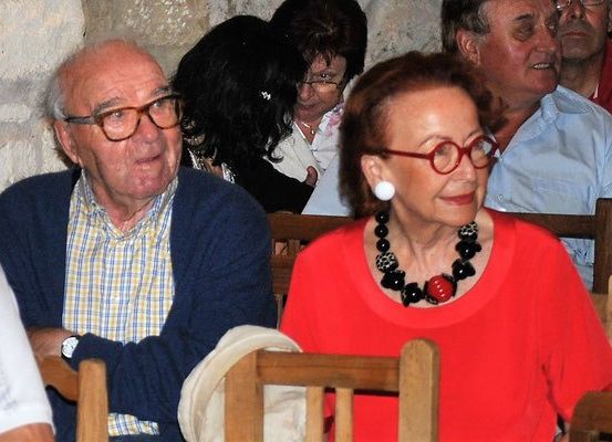Ein älteres Paar sitzt inmitten einer Gruppe von Menschen, beide mit Brille und roten Kleidungsstücken. Der Mann trägt eine Uhr, und die Frau trägt eine schwarze und weiße Halskette. Sie sitzen auf hölzernen Stühlen in einem Raum.