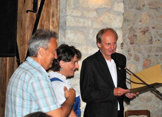 Ein Mann spricht in ein Mikrofon, während zwei andere hinter ihm stehen, einer in traditioneller Kleidung. Ein Lautsprecher ist an der Wand montiert.