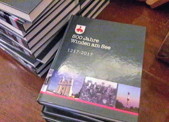 Ein Stapel Bücher mit dem Titel '800 Jahre Winden am See 1217-2017' auf einem Holztisch mit einem braunen Papierbeutel in der Nähe.
