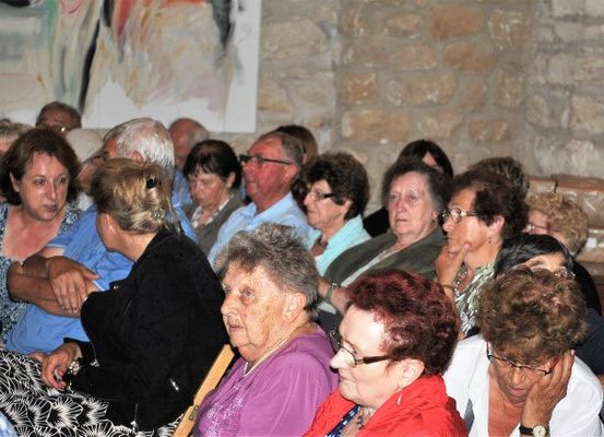 Eine Gruppe älterer Menschen sitzt in Stühlen und beobachtet eine Veranstaltung. Einige tragen Brillen und Halsketten. Hinter ihnen steht eine Steinmauer.