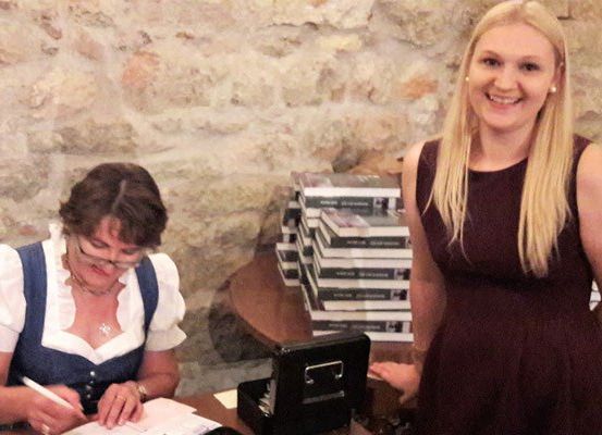 Eine Frau in traditioneller Kleidung schreibt auf ein Stück Papier. Eine lächelnde Frau in einem Kleid steht neben ihr. Dahinter befindet sich eine Steinmauer und ein Tisch mit Büchern.