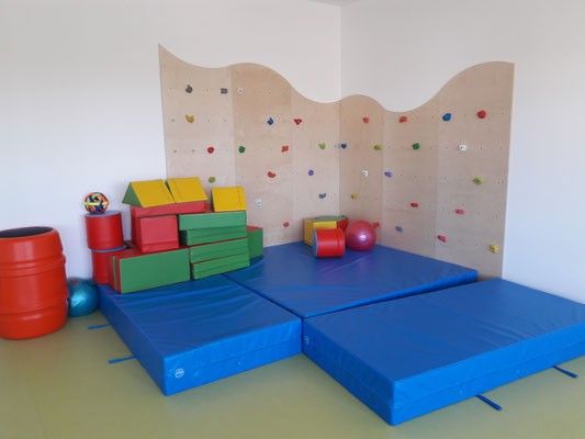 Ein Spielbereich mit blauen Matten, bunten Bauklötzen, einem roten Fass und einer Kletterwand mit verschiedenen Griffen.