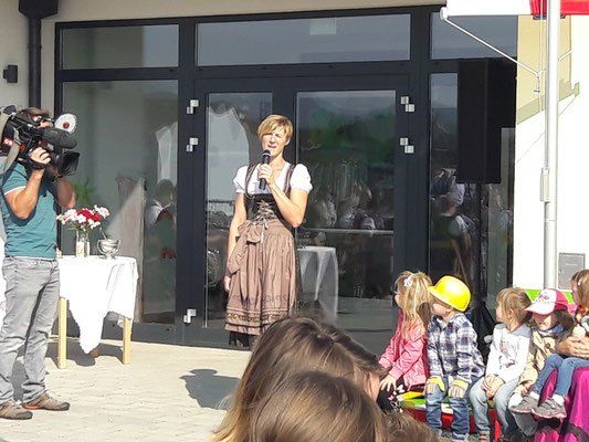 Eine Frau steht auf der Bühne, hält ein Mikrofon und scheint zu einer Gruppe von Kindern und Erwachsenen zu sprechen. Ein Mann steht hinter ihr und hält eine Videokamera. Links steht ein Tisch mit Blumen.