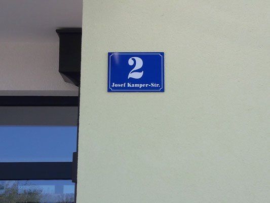 Ein blaues Schild mit der Nummer 2 und 'Josef Kamper-Str.' befindet sich an einer weißen Wand. Die Wand befindet sich neben einem Fenster.