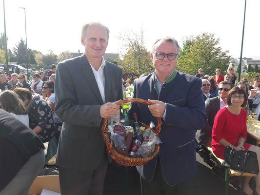 Zwei Männer stehen mit einem Korb voller Produkte auf einem öffentlichen Außengelände, mit einer Menge von Menschen um sie herum.