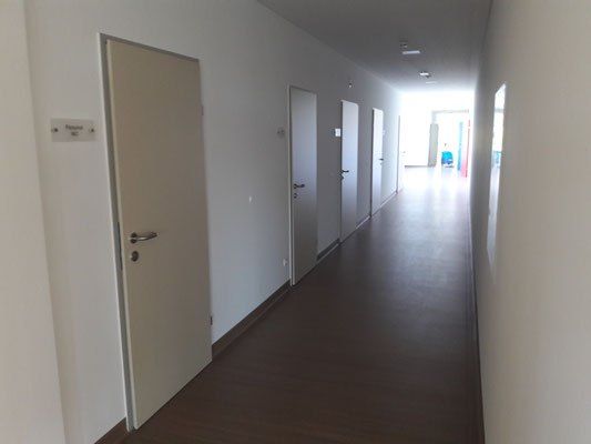 Ein leerer Flur mit weißen Wänden, Holzböden und mehreren geschlossenen Türen auf beiden Seiten. Es gibt ein kleines Schild an der Wand links.