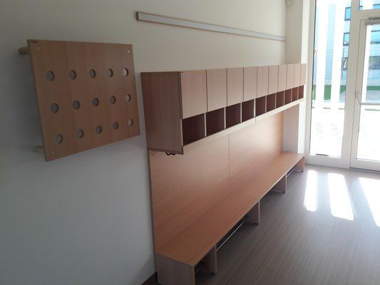 Ein leerer Raum mit Holzmöbeln, darunter eine Bank, Schränke und eine Lochtafel an der Wand. Sonnenlicht strömt durch Glastüren.